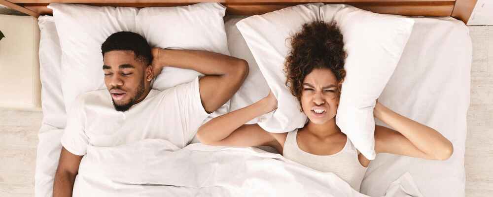 Man sleeping and snoring with sleep apnea while his wife is trying to block out the noise with her pillow